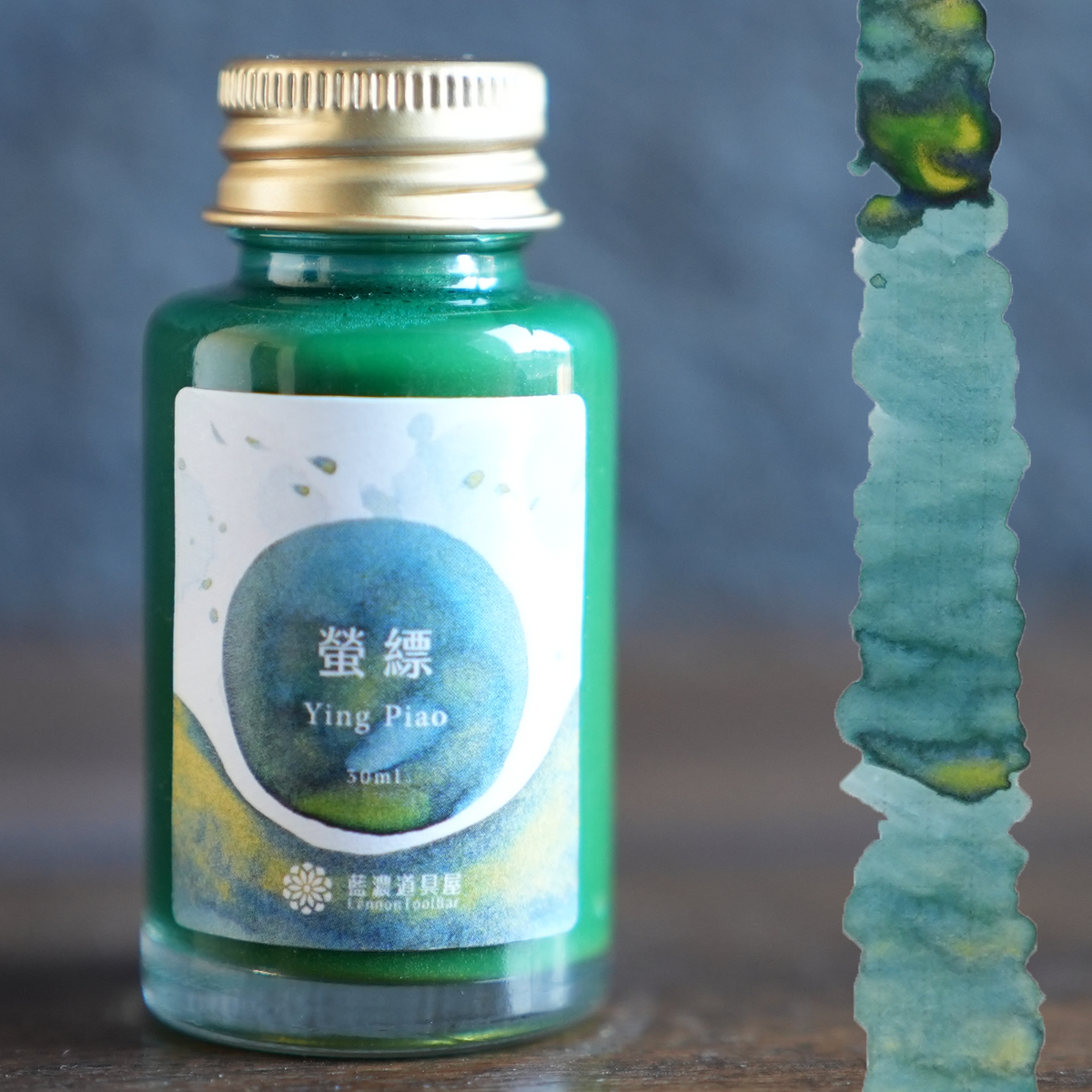 Lennon Tool Bar Ying Piao bottle on a wooden desk next to a swatch for it.  A green ink with yellow shading and hold and green shimmer. 